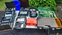 Wide view of all items on table including multiple tool cases, electric sander, green garden tool set, gloves, and painting supplies.