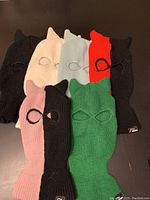 Photo of six knit full-face knit face masks in different colors with cat ears detail, laid out on a flat surface.