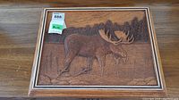Full view of framed wooden moose carving showing the animal in a natural wetland environment with trees in the background.
