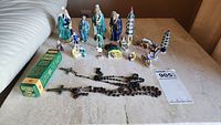 Photo of the entire lot showing multiple Asian pottery figurines including people in traditional attire, pagoda structures, and two rosaries with crucifixes on a beige surface.