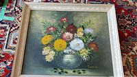Framed oil painting on board showing a bouquet of colorful flowers in a decorative vase with a muted background.