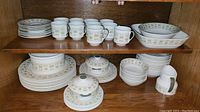 Full display of dinnerware set including stacked plates, cups, bowls, lidded bowls, and creamer on wooden shelves.