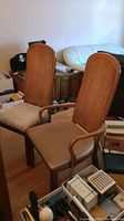 Two captain's chairs side by side on wood floor with beige seats and cane backs, surrounded by other household items and furniture