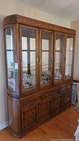 Front view of Sklar Peppler wood and glass lighted display hutch with glass doors, wooden base cabinetry, and multiple glass shelves inside.