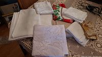Tablecloths, placemats, and napkins folded and placed on a lace covered table, showing range of fabric textures and colors.