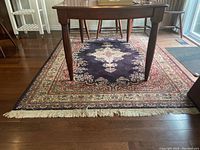 Area rug shown under a wooden dining table on hardwood floor. Dark blue field with ornate floral border and cream/red central medallion visible. Fringed tassels on one edge shown.