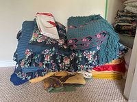 Pile of bedding and blankets including floral comforters, blankets, and thick quilted bed linens stacked on floor