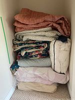 Stack of assorted blankets and bedding including a pink blanket on top, white with blue small floral print underneath, multicolored floral patterned fabric, and pale pink and beige bedding at the bottom.