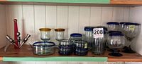 Wide shot showing assortment of blown glass pieces on shelf including goblets, drinking glasses, covered dish, and wine stoppers.