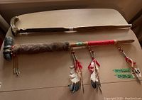 Full view of both items on a flat surface showing the carved wooden cane and the peace pipe with fur and feather decorations.