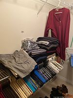 Wide view of shelf with multiple clothing items including shorts, folded sweaters, shirts, and hangers. Maroon long sleeve shirt hanging on the right side.