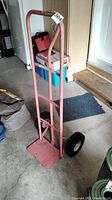 Full view of pink metal dolly hand truck standing upright on garage floor with handle and wheels visible.