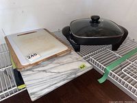 Shelf showing black West Bend electric skillet with glass lid beside three cutting boards: white plastic, wooden with handle, and large stone with marble look.