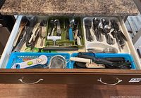 Kitchen drawer open showing flatware including forks, spoons, knives, and various kitchen tools like peelers, a digital thermometer, and serving utensils organized in trays and compartments.