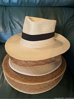 Four stacked woven straw hats in assorted natural colors and different weave patterns with black ribbon bands on three.