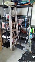 Wooden folding ladder leaning against shelving with visible paint wear and splatter, next to metal step ladder.