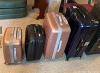 Four hard-sided suitcases lined up side-by-side on carpet floor: two rose gold, one black, one large blue suitcase.