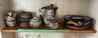 Shelf view of all pottery items showing cups, plates, creamers, a sugar bowl, a tea pot, and a large serving bowl with floral pattern.