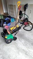 Photo of green electric snow blower with yellow and red handled shovels leaning against it on a concrete floor in a garage.