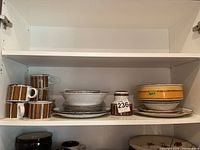 Stonehenge Midwinter mugs with brown vertical stripes stacked beside plates and serving bowls in brown and beige tones.