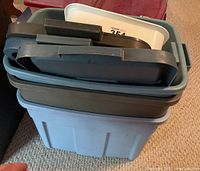 Stack of four large blue-gray plastic storage totes with lids and one smaller white plastic tote on top on beige carpet