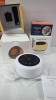 White noise machines displayed with their boxes: ColourNoise model visible with user manual and circular device with black control panel.