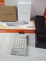 Photo of external CD/DVD drive box, white mini keydial box, Newmen GM610 keyboard box, and Satechi wireless keypad box arranged on table.