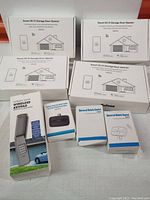 Photo showing four boxes of Smart Wi-Fi Garage Door Openers RSG100, one box of wireless garage door keypad, and two boxes of universal remote controls SM24, all in factory packaging, arranged on a flat surface.
