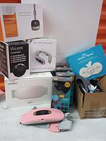 Full view of all items in lot including boxed neck massagers, vibrating massage ball, Orb Kuaplan trackers, and wearable pink massager