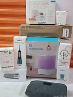 Photo showing the full lot of personal wellness items stacked together against an orange background, including boxes for humidifier, heating pad, oral irrigator, manicure set, massager, and digital scale.