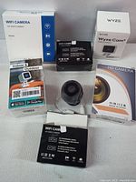 6 WiFi cameras in factory packaging including Wyze Cam and generic models boxed with accessories on white backdrop