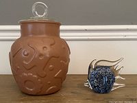 Front view of brown glass cookie jar with abstract embossed patterns and a clear glass lid with round handle next to the blue millefiori glass fish paperweight.