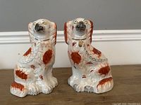 Pair of white ceramic Staffordshire spaniels with orange-brown markings and gold collars shown together on wooden surface against gray background.