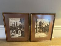 Pair of two framed watercolour paintings with brown mats and goldish wooden frames, leaning against a grey wall.