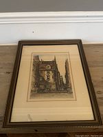 Full view of framed antique coloured etching lying flat against a baseboard and floor, showing the entire frame and visible artwork inside.