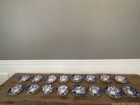 Overview of all 16 Royal Crown Derby Blue Mikado saucers arranged on a wooden surface.