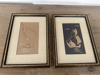 Two framed artworks side by side on a wooden surface, each depicting a boat made from natural materials on colored backgrounds.