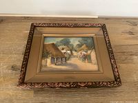 Framed watercolour painting of a 19th century village scene with thatched cottages and trees, view of entire framed artwork on a wooden surface.