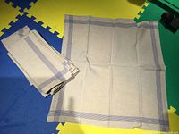 Ten folded blue and grey linen dinner napkins neatly stacked beside one napkin unfolded to show size and pattern.