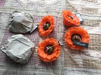 Six vibrant orange flower-shaped floating candles by Pont A La Ligne, three shown unpackaged, three wrapped in brown paper on a quilted bedspread.