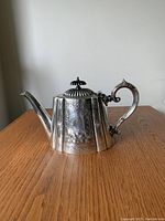 Front view of silver plated antique teapot showing ornate engraved design, curved spout and handle with thumb rest, and detailed domed lid with finial.