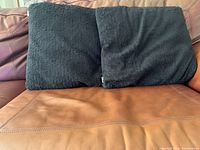 Front side view showing two black fuzzy cushions placed on a brown couch, showcasing their size, texture and color.