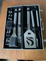 Full set of Cuisinart barbecue utensils and accessories organized inside open metal case on wooden surface.