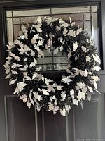 Full view of black plastic Halloween wreath with many small white ghost shapes covering it, hanging on a door with a glass window.
