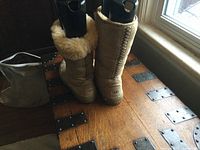 Front view of both beige UGG boots standing upright on a wooden trunk next to a window. Shows the exterior look and sole thickness.
