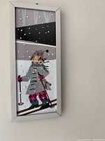 Front view of original framed picture of skier showing fabric and painted art, gray frame, and snow detail.