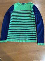 Full front view of long sleeve women's sweater laid flat showing green base with navy blue stripes and solid navy sleeves with gold buttons at shoulder.