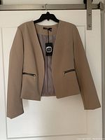 Full front view of the beige blazer with zippered pockets, hanging on a black hanger.