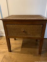 Front view of small wooden accent side table with woven wicker drawer front and knob handle, standing on a wood floor.