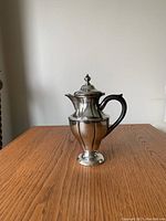 Front view of the vintage silver plated coffee pot showing the body, lid, spout, and black handle on a wooden surface.